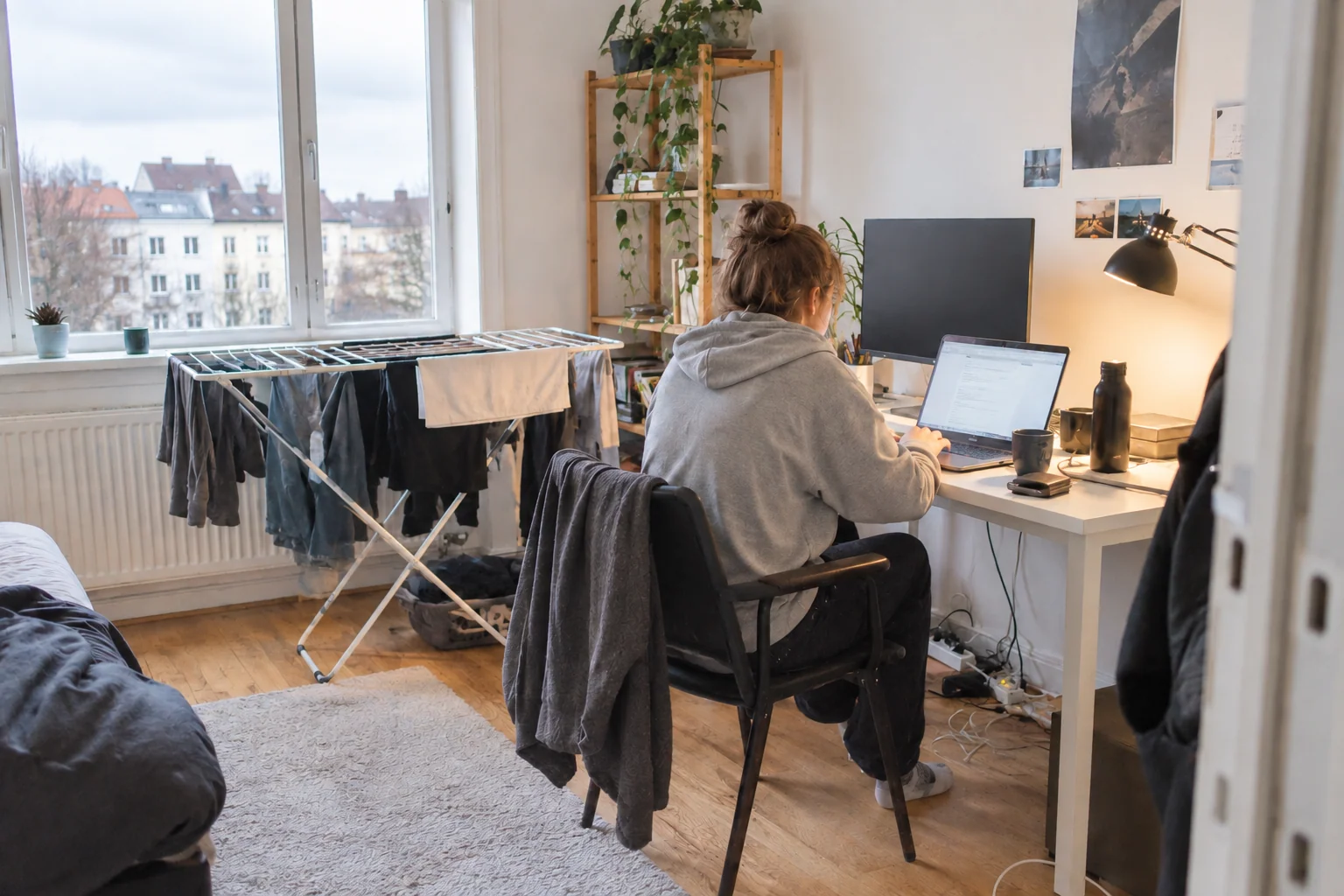 Person i hættetrøje arbejder ved et lille skrivebord med vasketøj i baggrunden